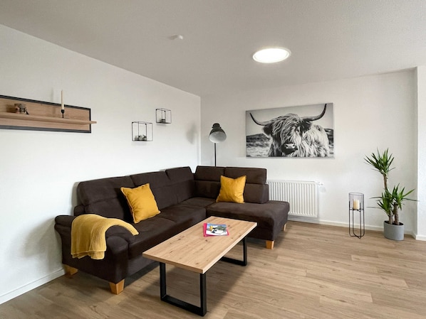 En moderne stue med en mørkebrun sofa, et træsofabord og en gulvlampe i sort. Et sort-hvidt fotografi af en tyr hænger på væggen, og der står en lille potteplante og et radiator.