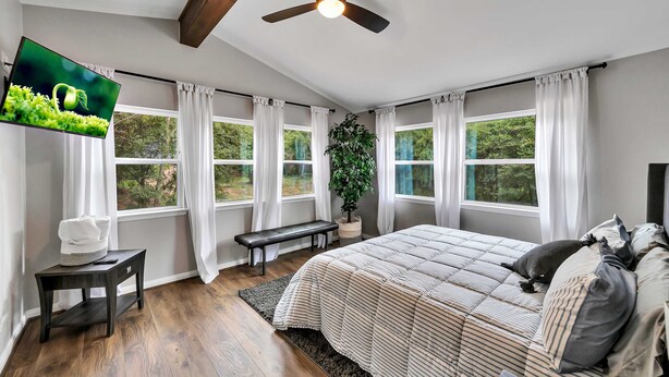 A bedroom with a bed, a ceiling fan, a TV, a bench, and a window with curtains. - Brazos River Bungalow - Close To Silos & Baylor (Waco)