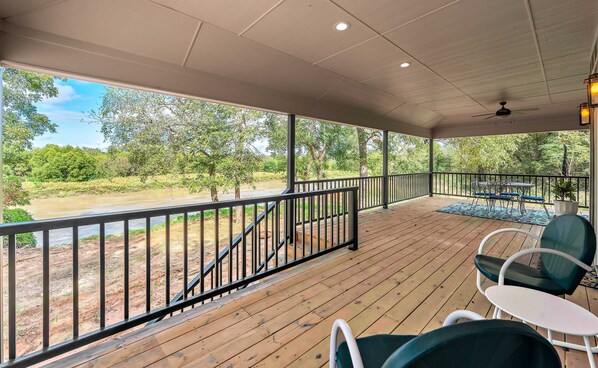 A covered outdoor area with a wooden deck, metal railing, and a view of a river and greenery. - Brazos River Bungalow - Close To Silos & Baylor (Waco)