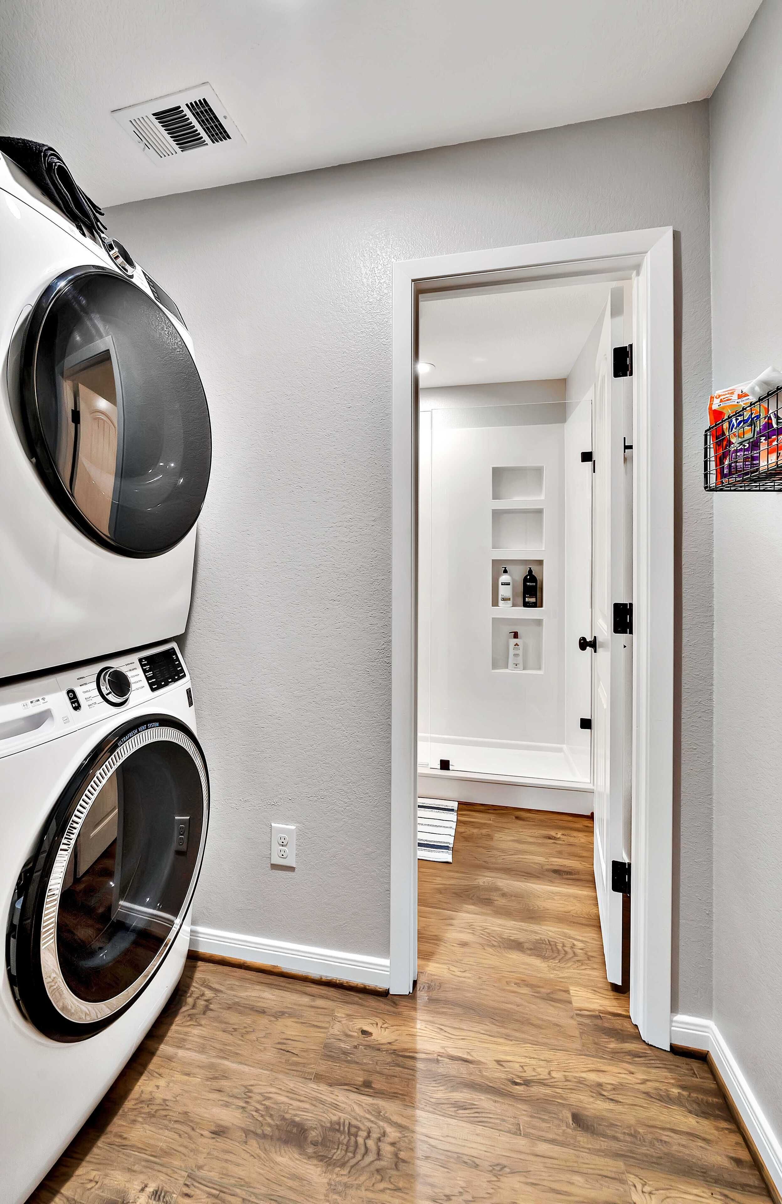 Une buanderie équipée d’un lave-linge et d’un sèche-linge à chargement frontal, un couloir menant à une pièce dotée d’une porte, et une étagère avec des bouteilles.