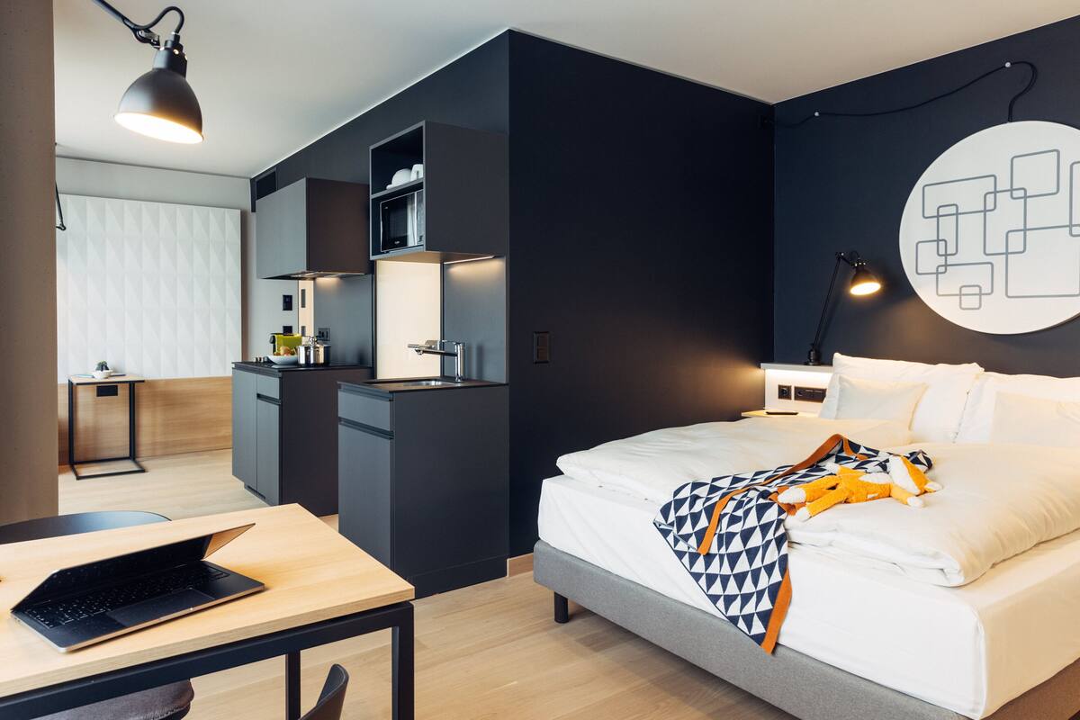 Une chambre d’hôtel moderne équipée d’un lit, d’un bureau avec un ordinateur portable, d’une kitchenette et d’un luminaire fixé au mur.