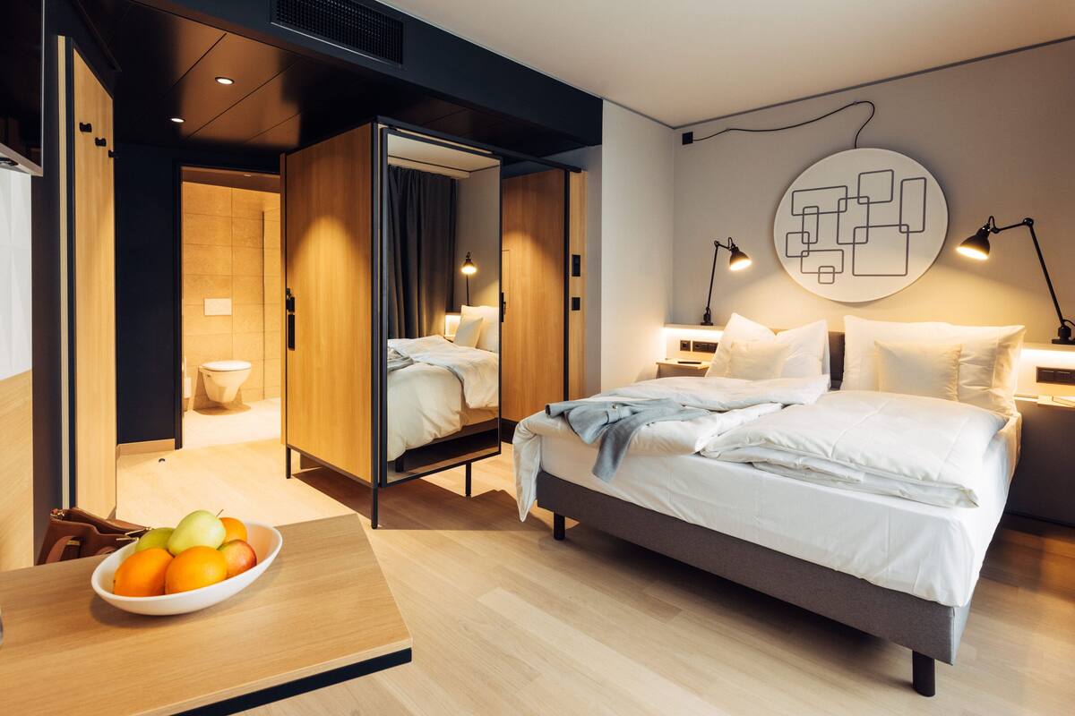 Une chambre d’hôtel moderne dotée d’un grand lit, de tables de chevet, d’un panier de fruits et d’une œuvre d’art murale originale.