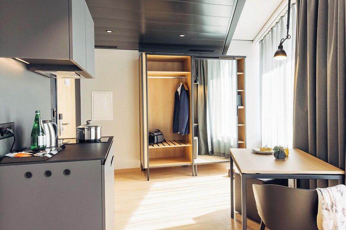 A modern hotel room with a kitchenette, a wardrobe with hanging space, a dining area, and a window with curtains.