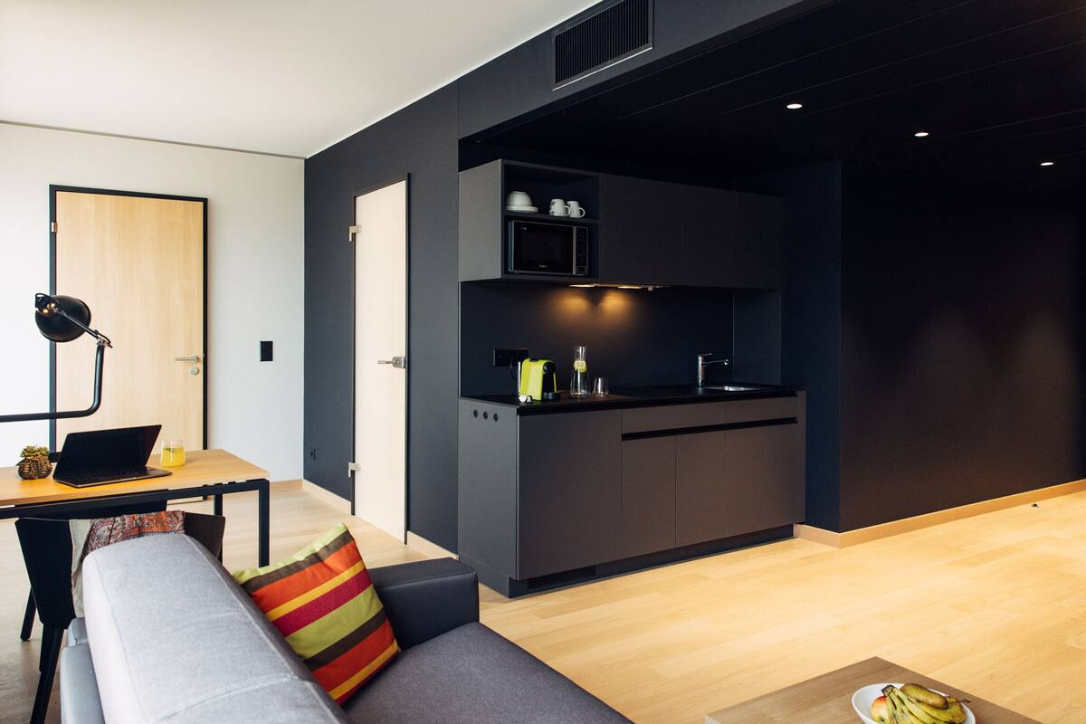 Une cuisine moderne avec des meubles de couleur sombre, un four à micro-ondes et un évier. On y trouve également un coin repas avec une table et un ordinateur portable.