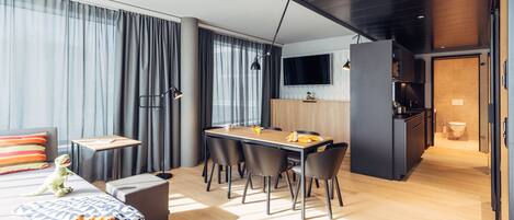 Habitación de hotel moderna con comedor, sofá, una mesita con lámpara y televisor montado en la pared.