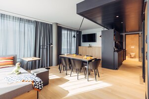 Habitación de hotel moderna con comedor, sofá, una mesita con lámpara y televisor montado en la pared.