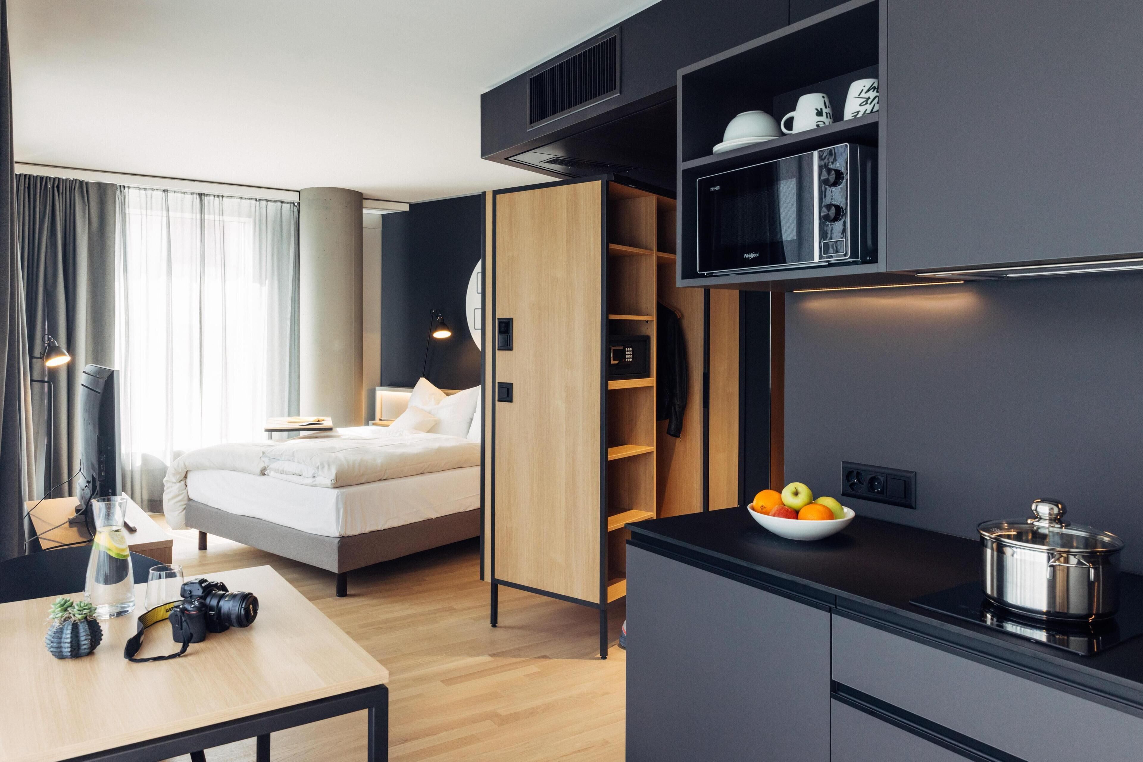 A modern hotel room with a bed, a desk with a camera, a bowl of fruit, and a microwave.
