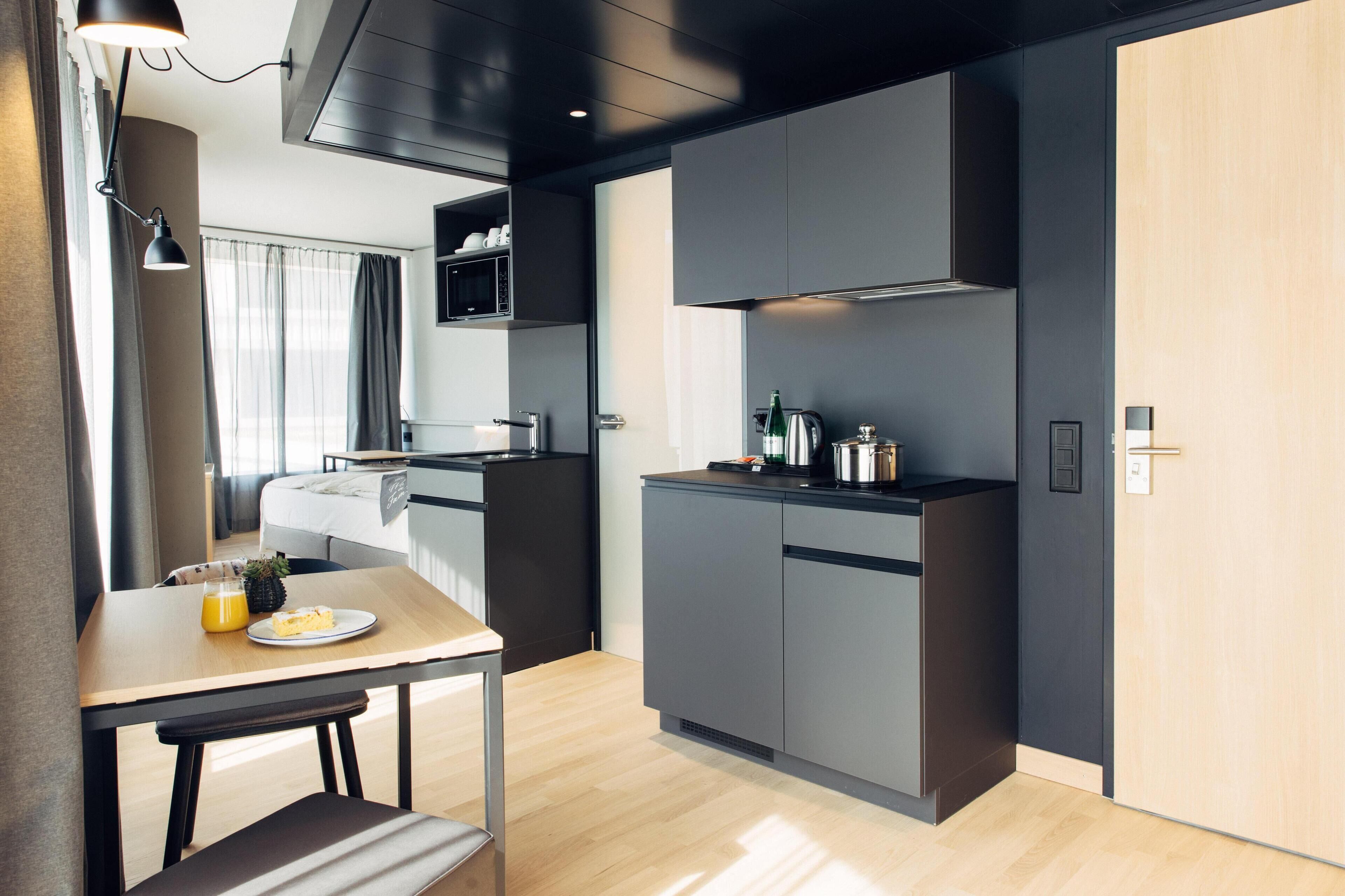 Una cocina compacta con electrodomésticos modernos, un comedor con mesa y banco, y una cama visible al fondo.
