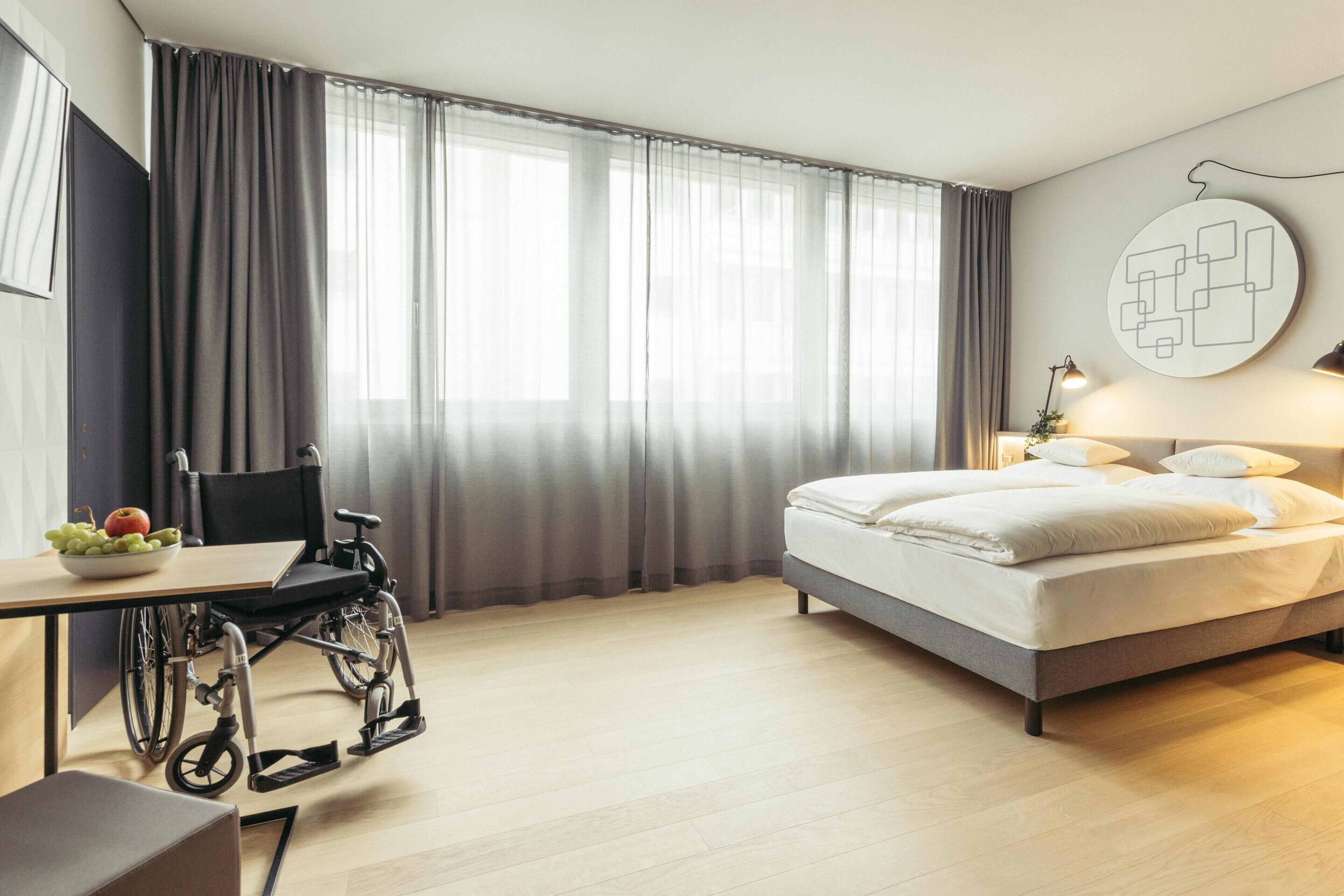 A hotel room with a bed, a desk, a wheelchair, and a large window with curtains.