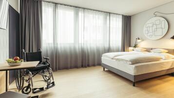 A hotel room with a bed, a desk, a wheelchair, and a large window with curtains.