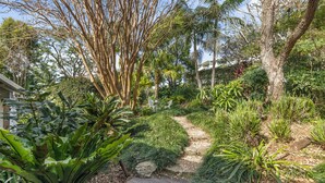 A garden pathway with various plants and trees, leading to a building with a white exterior.