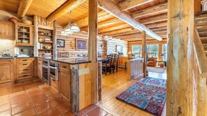 A rustic cabin interior with wooden beams, a kitchen with wooden cabinets, a dining area, and a large rug. - 88 Potentilla Lane by Summit County Mountain Retreats (Keystone)