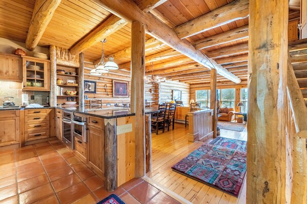 A rustic cabin interior with wooden beams, a kitchen with wooden cabinets, a dining area, and a living room with a large window. - 88 Potentilla Lane by Summit County Mountain Retreats (Keystone)