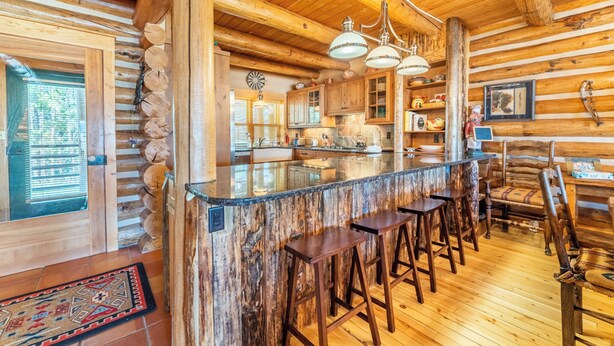 A rustic log cabin interior with a central bar area, wooden flooring, and a high ceiling with exposed beams. - 88 Potentilla Lane by Summit County Mountain Retreats (Keystone)