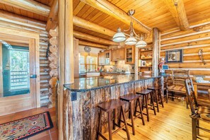 A rustic log cabin interior with a central bar area, wooden flooring, and a high ceiling with exposed beams. - 88 Potentilla Lane by Summit County Mountain Retreats (Keystone)