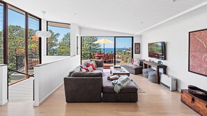 A spacious living room with a large sectional sofa, a coffee table, and a TV. There is a balcony with a view of the ocean and trees. - Magnificent 3BR Bronte Holiday House with Views (Bronte)