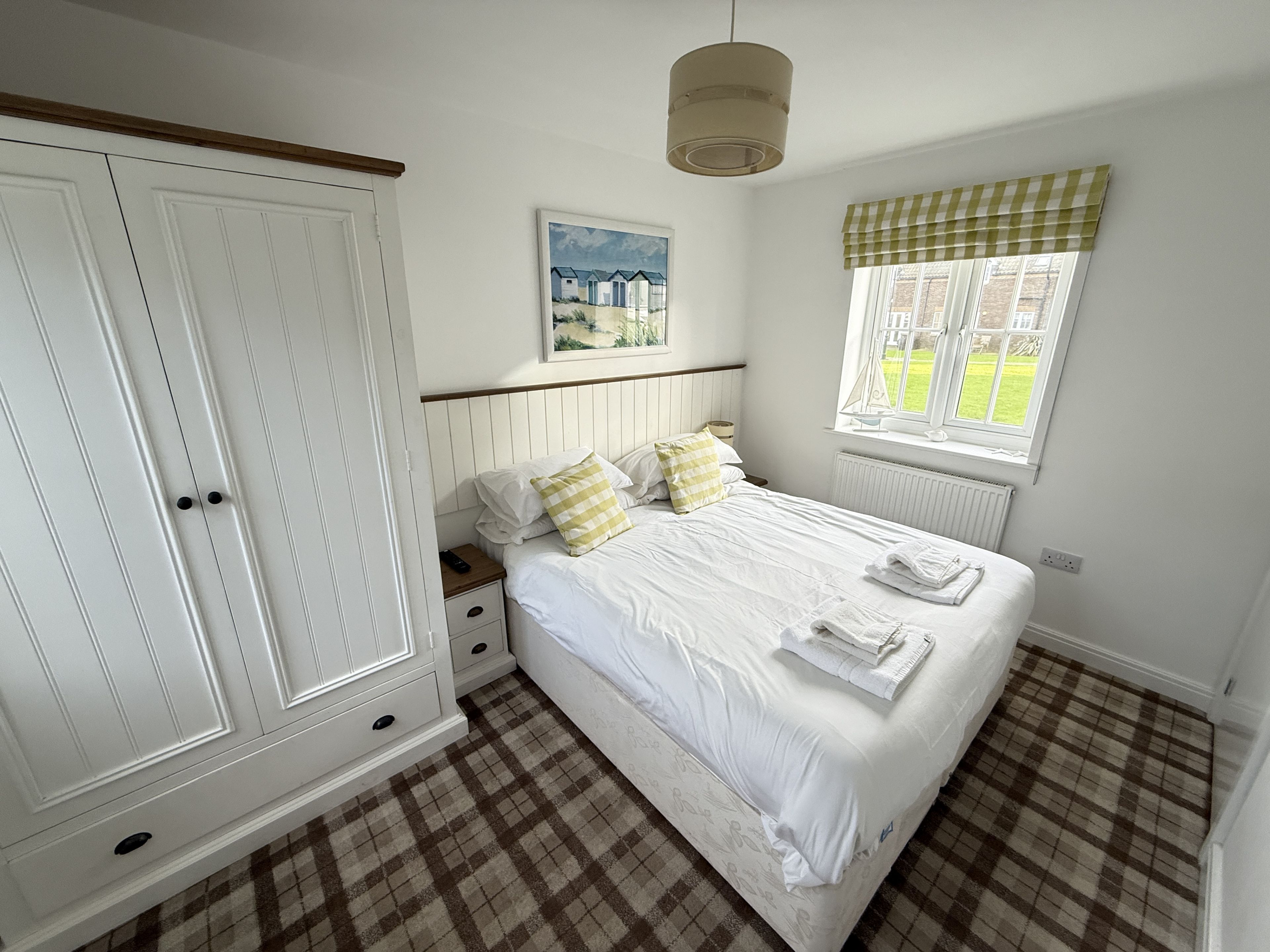 A bedroom with a large wardrobe, a bed with white linens, a checkered floor, a window with a view, and a painting on the wall.