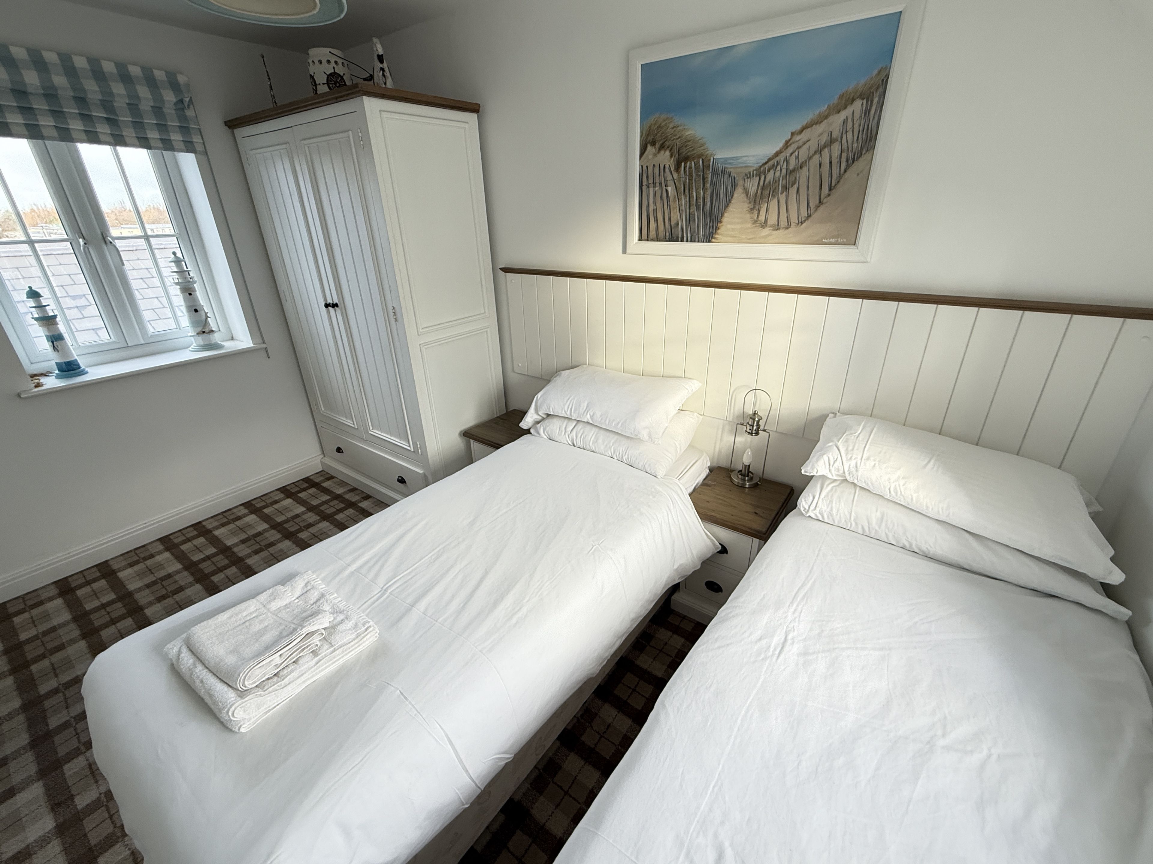 A hotel room with two single beds, a wardrobe, a painting of a beach, and a window with curtains.