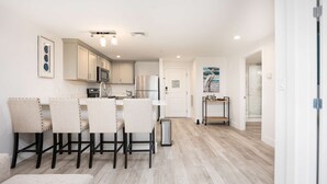 A modern kitchen with white cabinetry, a built-in microwave, and a stainless steel refrigerator. There is a dining area with four chairs and a small table. The floor is wooden, and there is a door leading to a bathroom with a glass shower. - Premier Ocean View Corner Condo (850S.F.) (Plymouth)