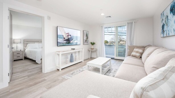 A modern living room with a large sofa, a coffee table, and a TV mounted on the wall. There is a balcony with a view of the sea. - Premier Ocean View Corner Condo (850S.F.) (Plymouth)