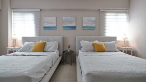 A hotel room with two beds, each with a white bedspread and a single yellow pillow. There are three paintings on the wall depicting a beach scene.