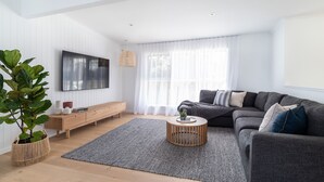 Ein modernes Wohnzimmer mit einem grauen Sofa, einem Holztisch, einem an der Wand montierten Fernseher und einem großen Fenster mit durchsichtigen Vorhängen.
