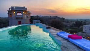 A swimming pool with a stone deck and lounge chairs, a small building with arched windows, and a sunset in the background. - Brij Lakshman Sagar, Pali (Raipur)