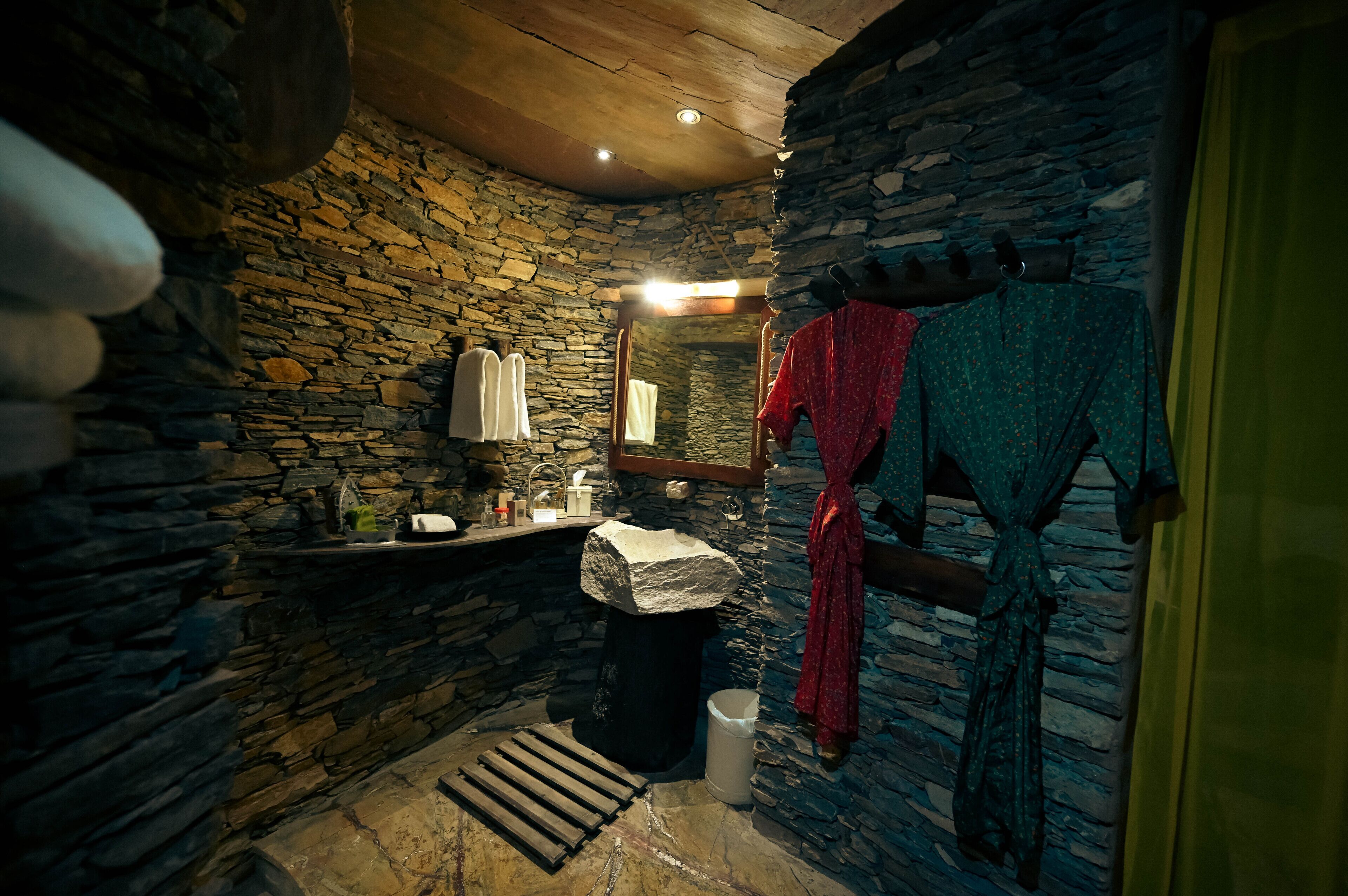 A stone-walled bathroom with a wooden ceiling, a sink, a mirror, and a towel rack with robes.