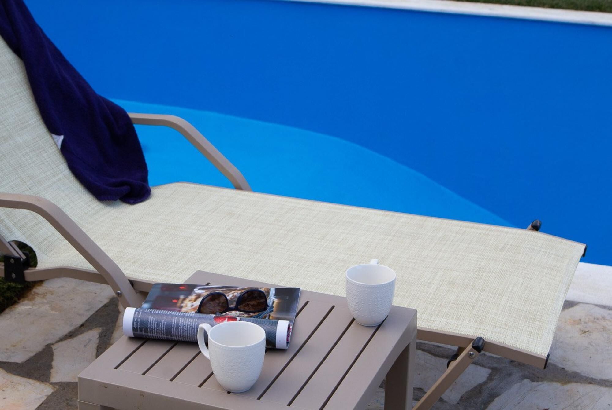 A poolside chair with a magazine, a white mug, and a glass of water.