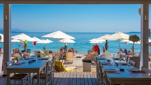 Zona d'estar a l'aire lliure amb taules i cadires, amb vista a una platja amb ombrel·les i gent.