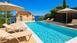 Una piscina amb gandules i ombrelles, un edifici amb teulada de rajola i vista al mar.