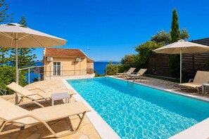 A swimming pool with lounge chairs and umbrellas, a building with a tiled roof, and a view of the sea.