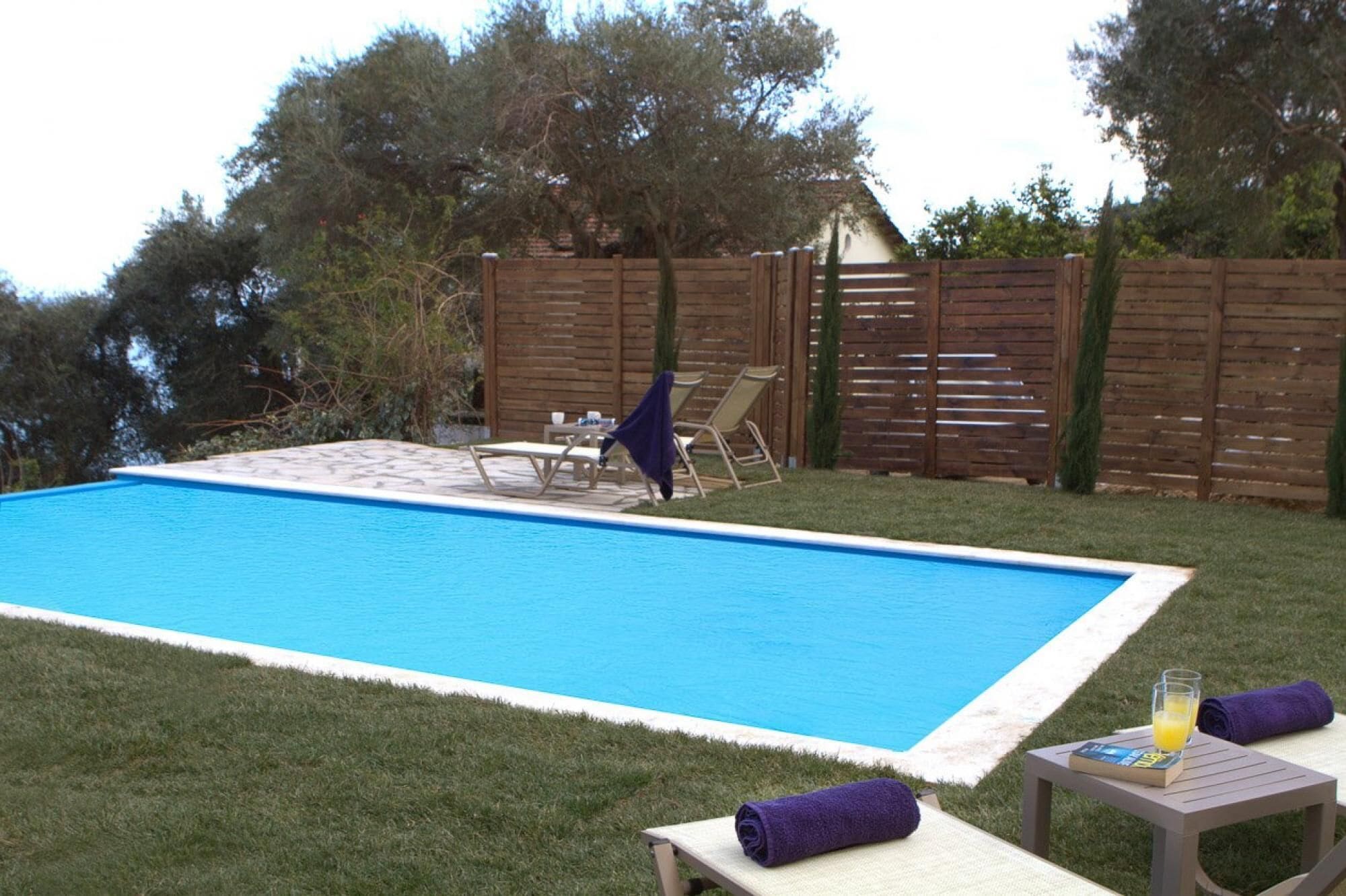 A swimming pool with a wooden deck, lounge chairs, and a table with a drink and a book.