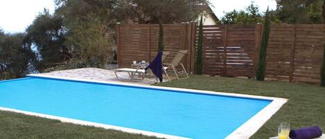 A swimming pool with a wooden deck, lounge chairs, and a table with a drink and a book.