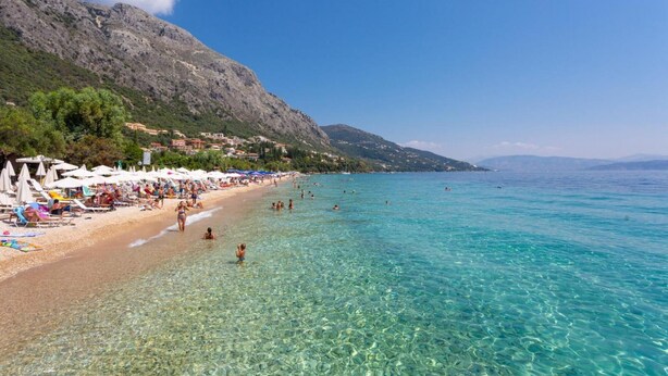 Una platja amb aigua turquesa cristal·lina, gent nedant i prenent el sol, i un paisatge de muntanyes al fons.
