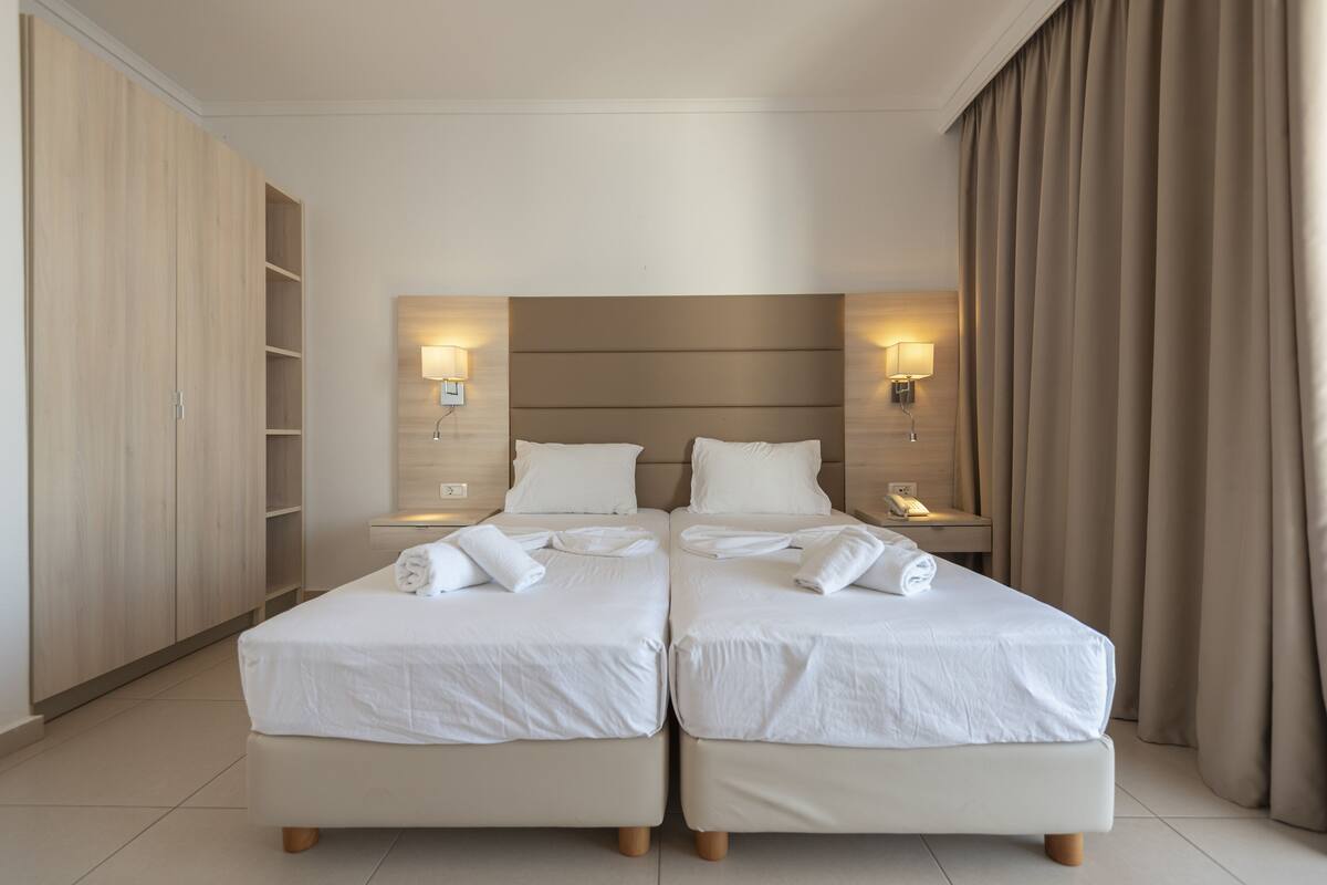Una habitación de hotel con dos camas, un armario, una mesita de noche con lámpara y una cortina.