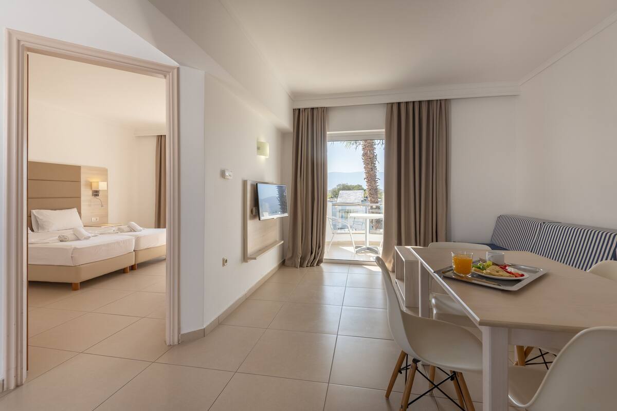 Habitación de hotel con cama, televisión, zona de comedor con mesa y sillas, y balcón con cortinas.