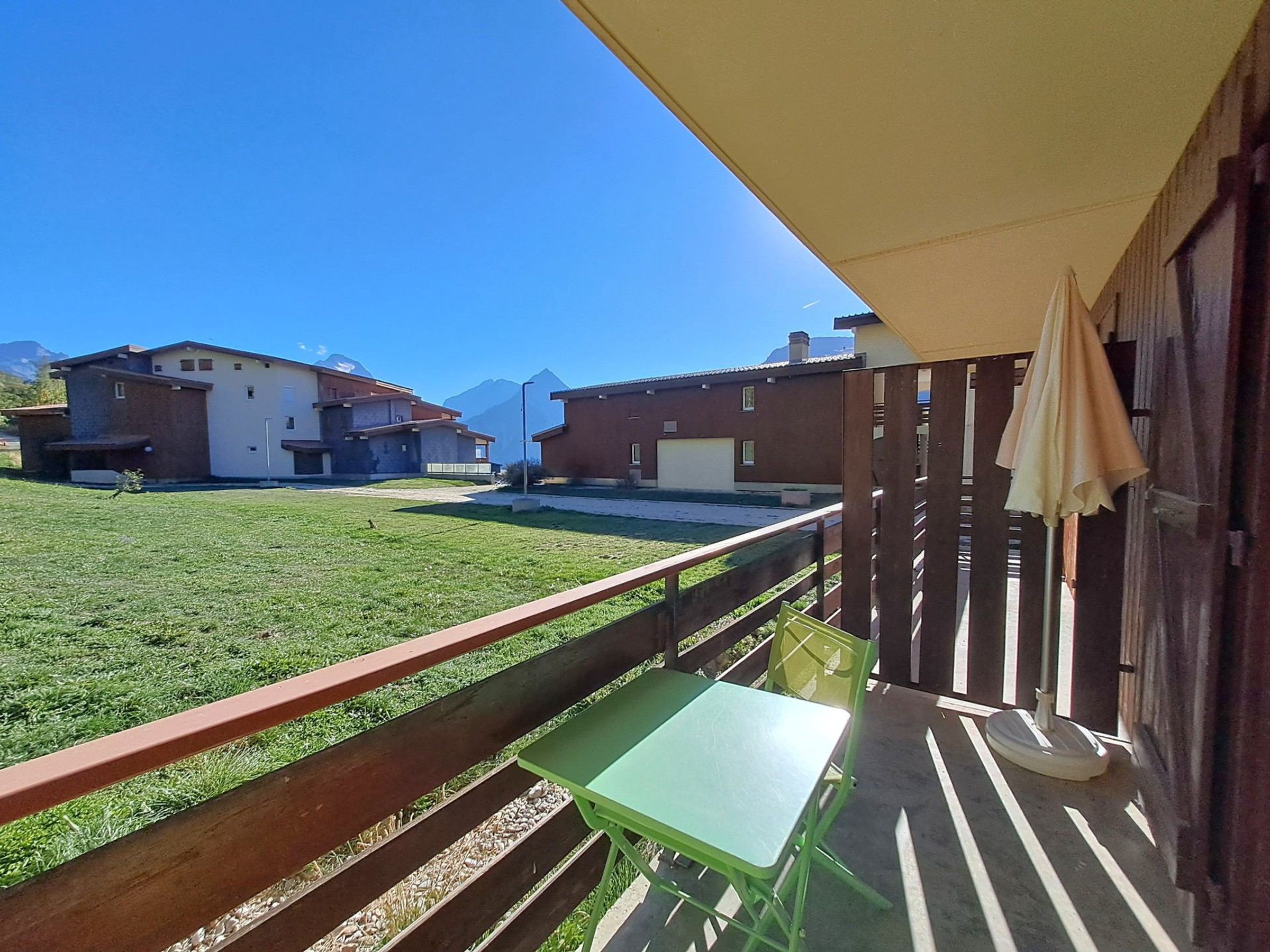 Uma varanda com mesa e cadeiras verdes, com vista para uma área gramada e edifícios ao fundo.