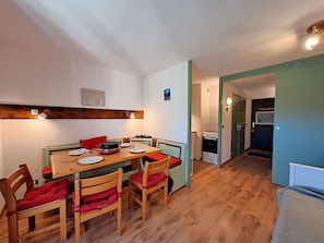 A dining area with a wooden table and chairs, a kitchen in the background, and a door leading to another room.