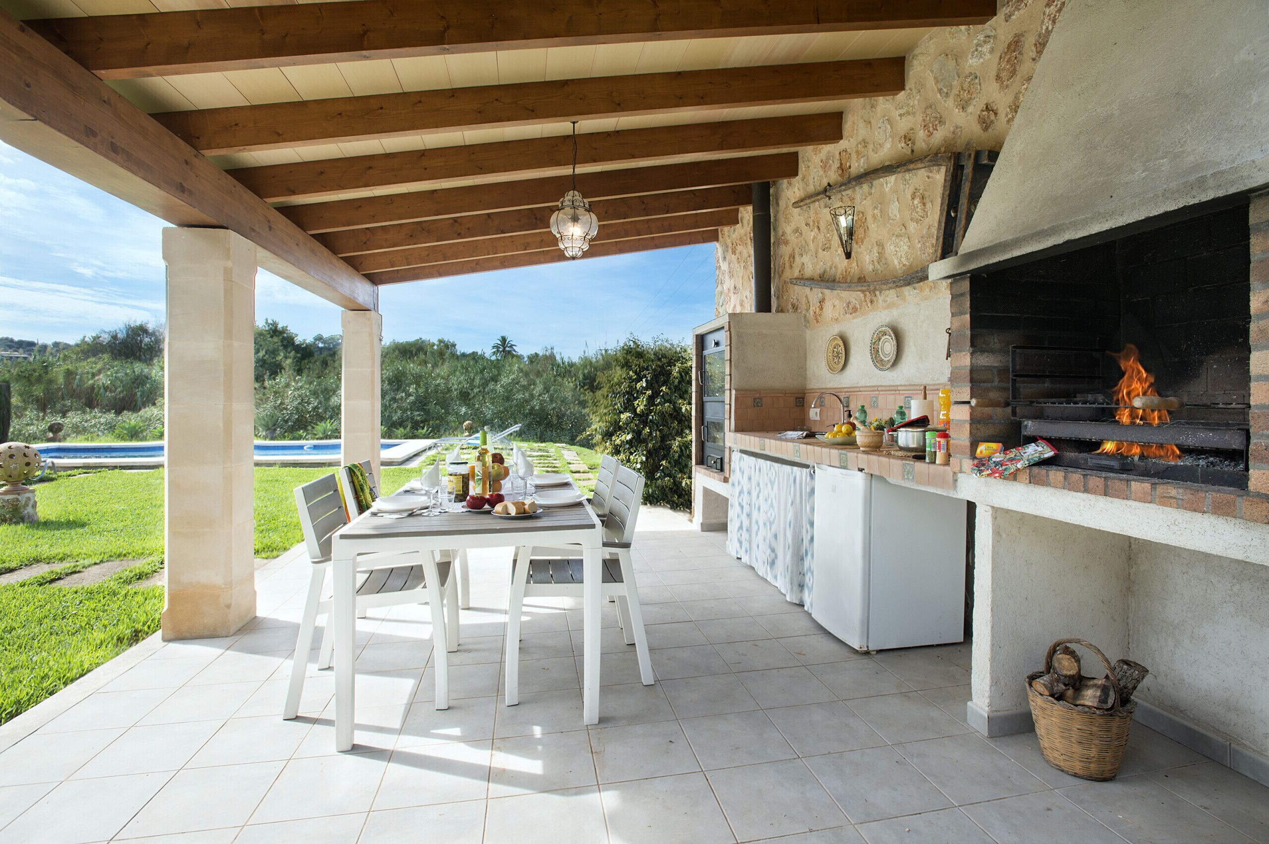 Cucina esterna con parete in pietra e camino in mattoni, tavolo da pranzo con sedie e vista su un giardino rigoglioso.
