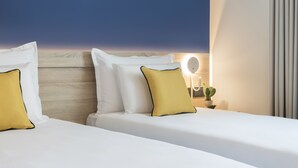 A hotel room with two beds, each with white bedding and yellow pillows, a wooden headboard, a bedside table with a lamp and a cactus, and a view of the sky. - Antoniel Suites (St. Julian's)