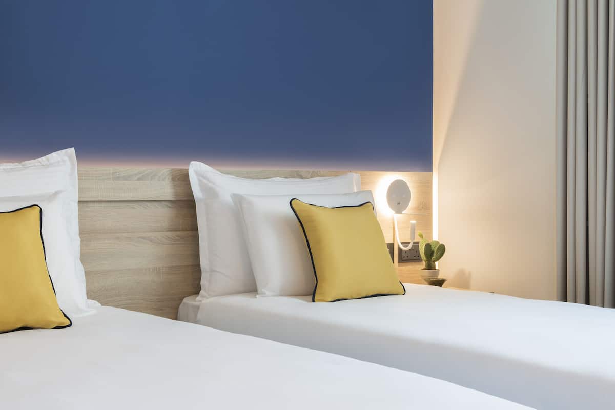A hotel room with two beds, each with white bedding and yellow pillows, a wooden headboard, a bedside table with a lamp and a cactus, and a view of the sky.