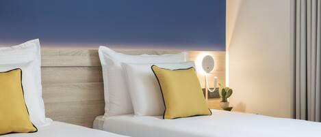 A hotel room with two beds, each with white bedding and yellow pillows, a wooden headboard, a bedside table with a lamp and a cactus, and a view of the sky.
