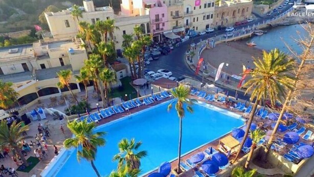A pool area with lounge chairs and palm trees, surrounded by buildings with a Mediterranean architectural style. - Antoniel Suites (St. Julian's)