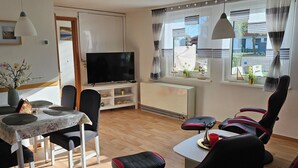 Ein gemütlicher Wohnraum mit Esstisch, Stühlen, Fernseher und einem Fenster mit Vorhängen.