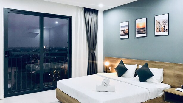 Une chambre d’hôtel moderne avec un grand lit, une table de chevet, une fenêtre donnant sur la ville et trois tableaux encadrés accrochés au mur.