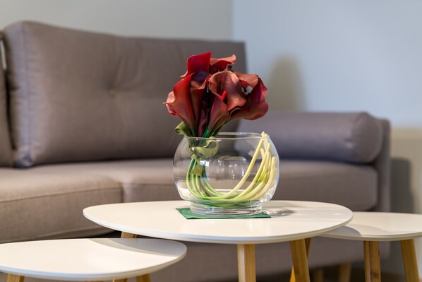 Un vase en verre contenant des fleurs, posé sur une table blanche dans un salon où se trouve un canapé.