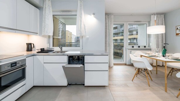 Eine moderne Küche mit weißen Schränken, einem eingebauten Ofen und einem Geschirrspüler. Ein Essbereich mit Tisch und Stühlen lädt ein, und ein großes Fenster bietet Blick auf die Stadt.
