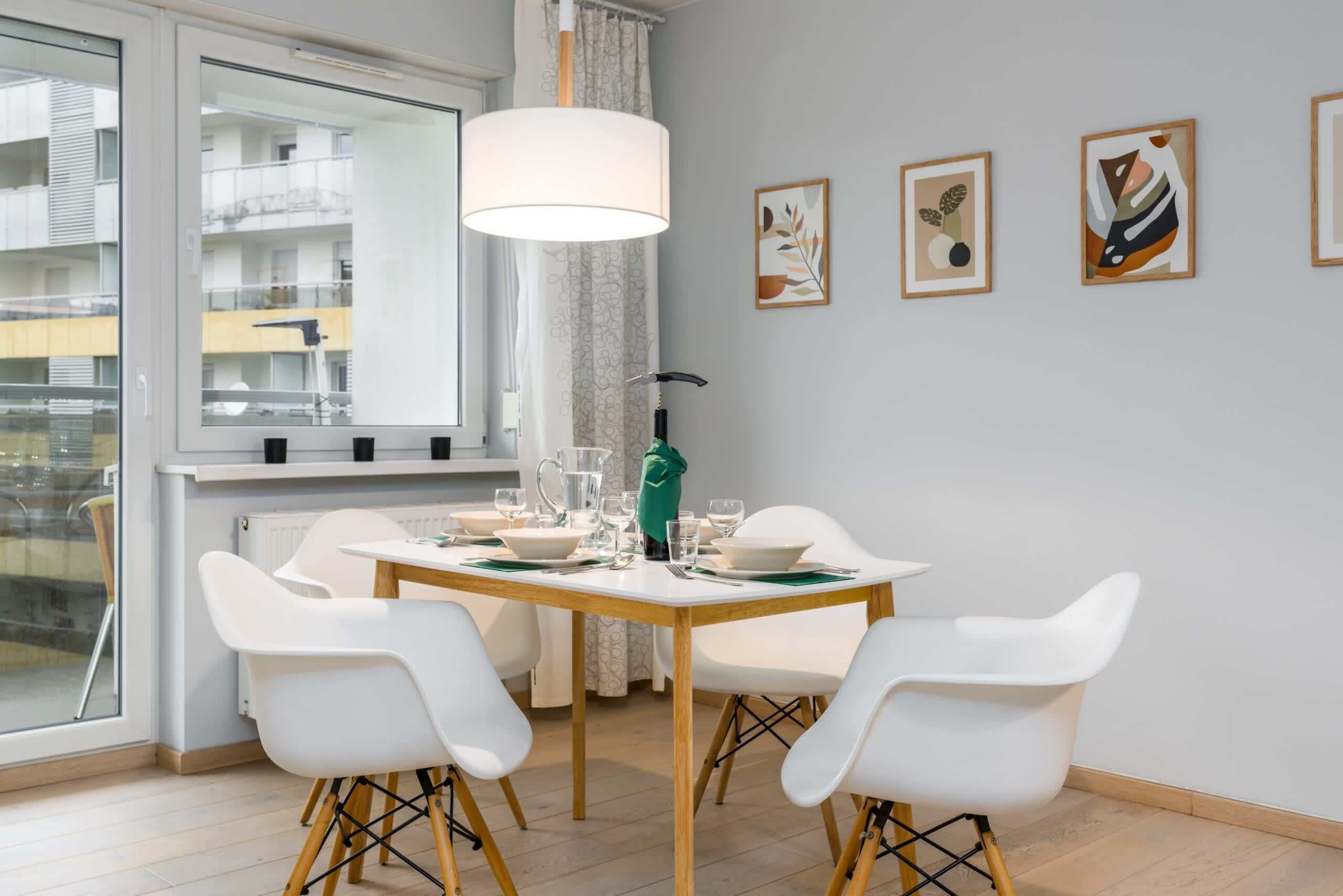 A dining area with a table set for four, white chairs, a hanging light fixture, and framed artwork on the wall.