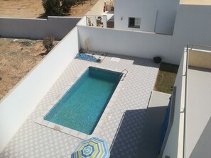 Una piscina en la azotea con solárium de baldosas, un pequeño jardín y barandilla metálica.